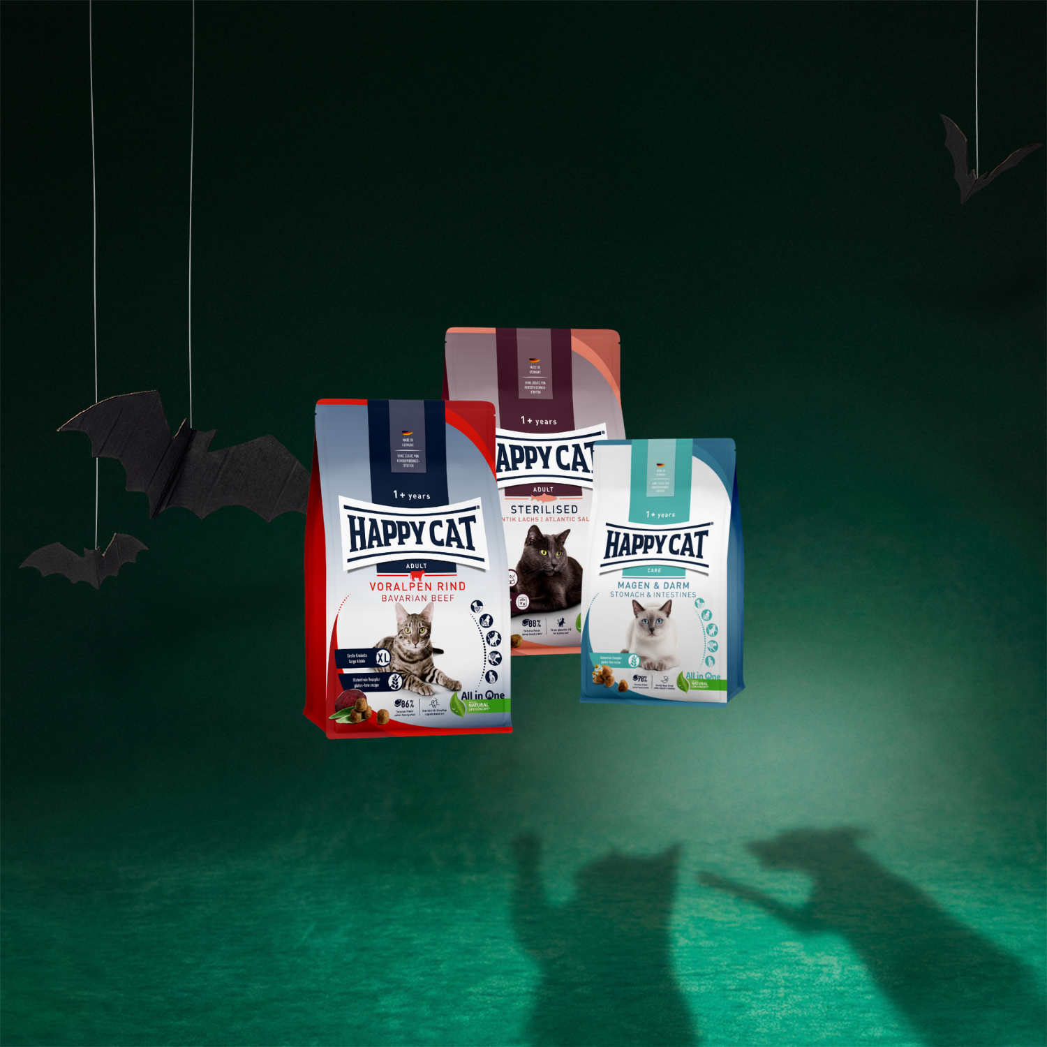 On voit ici trois paquets de croquettes pour chat de la marque Happy Cat, dans les variétés « Bœuf des Préalpes » (1,3 kg), « Saumon de l'Atlantique » (1,3 kg) et « Système gastro-intestinal » (300 g).