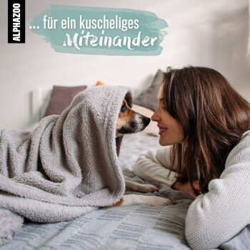 Ein Hund in einer grauen Decke liegt auf einem Bett und schaut auf eine Person in einem weißen Pullover. Textüberlagerung: "... für ein kuscheliges Miteinander."