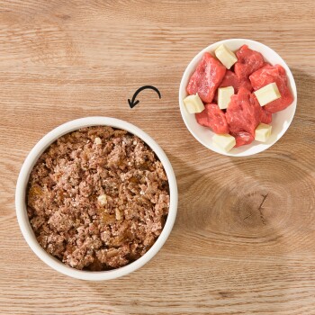 Eine Schüssel mit Haustierfutter, das eine texturierte, fleischige Mischung zeigt, steht links neben einer kleinen Schüssel mit rohen Fleischwürfeln und Fett.