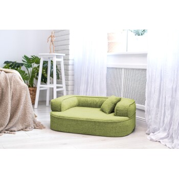 Ein gemütliches grünes Haustierbett mit einem kleinen Kissen, platziert in einem hellen Raum mit weißen Vorhängen und einer Topfpflanze im Hintergrund.