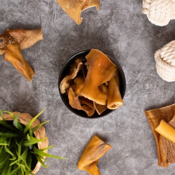 Eine schwarze Schüssel, gefüllt mit verschiedenen Hundekauartikeln, umgeben von verstreuten Kauartikeln und einem kleinen Korb mit grünem Gras, auf einer grauen Oberfläche.