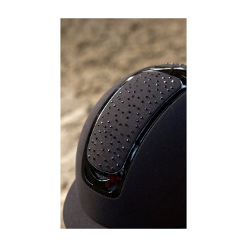 Nahaufnahme eines schwarzen Reithelms mit einer strukturierten, genoppten Oberseite und schlankem Design, vor einem verschwommenen sandigen Hintergrund.