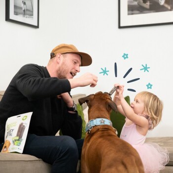 Un enfant en robe rose et un homme en pull noir partagent une friandise avec un chien marron, avec un sac de friandises pour animaux visible sur le canapé.
