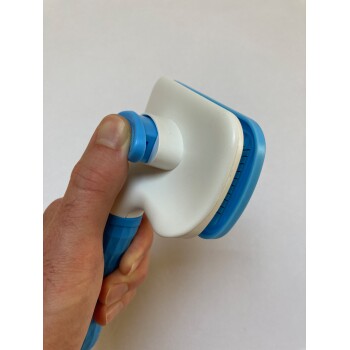 Eine Hand hält ein blau-weißes Haustierpflegewerkzeug mit einem strukturierten Griff und einem kammartigen Kopf, vor einem schlichten Hintergrund.