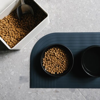 Ein weißer Behälter gefüllt mit trockenem Haustierfutter und einer Metallschaufel, neben einer schwarzen Schüssel mit mehr Trockenfutter auf einem dunklen Matten.