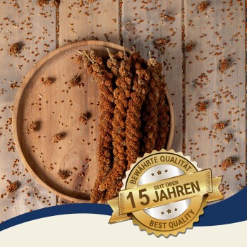 Getrocknete Hirseähren auf einem Holzbrett mit verstreuten Samen und einem Abzeichen mit der Aufschrift "Bewährte Qualität seit über 10 Jahren."
