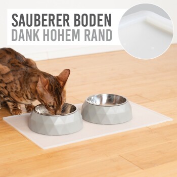 Eine Bengal-Katze frisst aus zwei grauen geometrischen Haustierschüsseln auf einer weißen Matte, mit dem Text 'SAUBERER BODEN DANK HOHEM RAND' darüber.