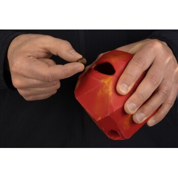Eine Person hält ein rotes, geometrisches Haustierspielzeug mit einem Loch und bereitet sich darauf vor, einen kleinen braunen Haustier-Leckerbissen einzufügen.
