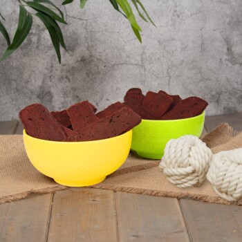 Eine schwarze Schüssel gefüllt mit rechteckigen Hundeleckerlis, umgeben von losen Leckerlis und weißen Seilspielzeugen, auf einer grauen Oberfläche.