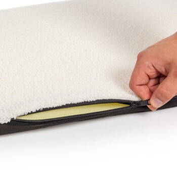 Eine Nahaufnahme eines Haustierbettes mit einer weichen weißen Fleece-Oberfläche und einem schwarzen Reißverschluss, der eine Hand zeigt, die den Bezug öffnet, um ein gelbes Innenfutter zu enthüllen.
