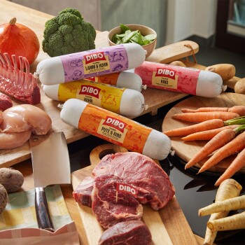 DIBO frische Tierfutterrollen mit den Aufdrucken Lamm & Huhn, Rind, Huhn, Pute & Rind mit rohem Fleisch, Gemüse und einem Fleischermesser auf einem Holztisch.