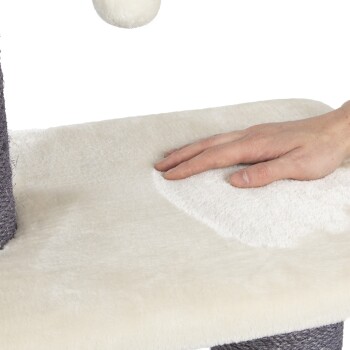 Eine Hand berührt sanft die weiche, cremefarbene Oberfläche eines Kratzbaums für Haustiere, der eine strukturierte graue Basis und einen hängenden weißen Pompon hat.