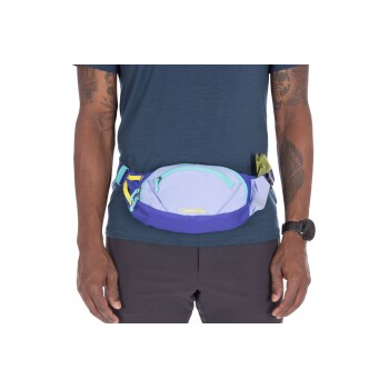 Eine Person in einem blauen Hemd und dunklen Hosen präsentiert einen bunten Ruffwear-Hüftgurt mit einem lila und blauen Design und einer grünen Tasche.