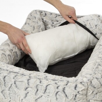 Eine Person legt ein weißes Kissen in ein plüschiges, graues Haustierbett mit strukturiertem Außenmaterial, das den weichen Stoff zeigt.