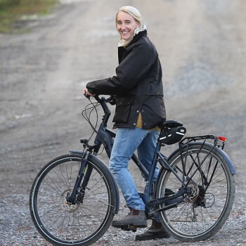 Person in schwarzer Jacke und Jeans steht mit einem schwarzen Fahrrad auf einem Kiesweg im Freien.