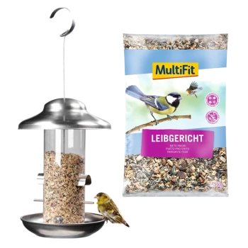 Mangeoire pour oiseaux remplie de graines, avec un petit oiseau perché sur la base ; un sac de nourriture pour oiseaux MultiFit étiqueté "Leibgericht" est à côté.