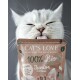 Eine Nahaufnahme einer weißen Katze, die sich die Lippen leckt, während sie ein Paket "CAT'S LOVE 100% Bio Junior" Katzenfutter hält, geeignet für 2-12 Monate alte Katzen.