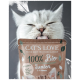 Eine Nahaufnahme einer weißen Katze, die sich die Lippen leckt, während sie ein Paket "CAT'S LOVE 100% Bio Junior" Katzenfutter hält, geeignet für 2-12 Monate alte Katzen.