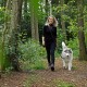 Eine Frau in schwarzer Kleidung führt einen flauschigen grau-weißen Hund an der Leine durch einen üppig grünen Waldweg.