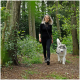 Eine Frau in schwarzer Kleidung führt einen flauschigen grau-weißen Hund an der Leine durch einen üppig grünen Waldweg.