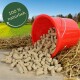 Roter Eimer, aus dem braune Tierpellets auf Holz und Stroh verschüttet sind, grüner Feldhintergrund; rundes Siegel mit der Aufschrift '100% natürlich'.