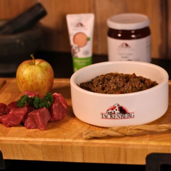 Biała miska karmy Tacrenberg dla psów z surowym mięsem, jabłkiem i pietruszką na drewnianej desce do krojenia.