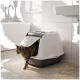 Chat tigré sortant d'une litière couverte blanche avec volet transparent sur un sol carrelé de salle de bain près d'un lavabo et d'une baignoire.