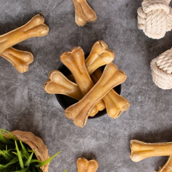 Eine schwarze Schüssel gefüllt mit mehreren Hundekau-Knochen, umgeben von einem Seilspielzeug und grünen Pflanzenakzenten auf einer grauen Oberfläche.