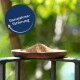 Ein Holzbrett hält einen Haufen Vogelfutter, mit einem verschwommenen grünen Hintergrund. Ein runder Aufkleber liest "Ganzjahresfütterung".
