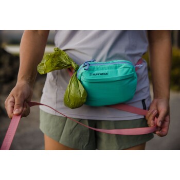 Eine Person hält eine rosa Leine in einer Hand und eine grüne Tasche, die an einem türkisfarbenen Ruffwear-Hüftgurt befestigt ist, in der anderen Hand, im Freien.
