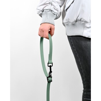 Eine Person hält eine mintgrüne Hundeleine mit einem schwarzen Clip, trägt eine hellgraue Jacke und dunkle Jeans, vor einem weißen Hintergrund.
