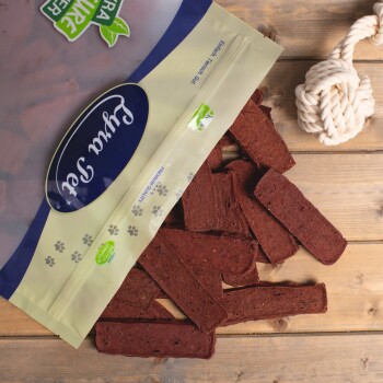 Eine Tüte "Premium Qualität" Haustierleckerlis kippt braune Leckerlis auf Holz, neben einem weißen Seilspielzeug.
