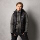 Eine Person, die eine schwarze Steppweste über einem grauen Reißverschluss-Hoodie trägt, steht selbstbewusst mit den Händen in den Taschen vor einem grauen Hintergrund.
