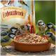 Kleine Singvögel, die geschälte Erdnussstücke aus einer Terrakottaschale fressen, im Hintergrund eine Tüte mit der Aufschrift 'Premium Erdnussbruch'.