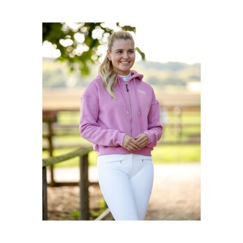 Eine Frau in einem rosa Reißverschluss-Hoodie steht im Freien, mit einem verschwommenen Hintergrund aus Grün und einem Holzzaun.