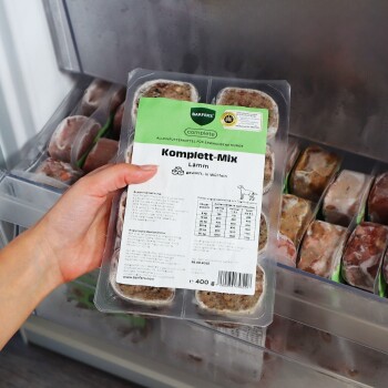 Hand hält gefrorene, verpackte Hundefutterportion ‚Komplett‑Mix Lamm‘ (BARFER'S Komplett), 400 g, im Gefrierschrank.
