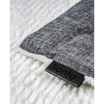 Nahaufnahme einer Ecke eines grauen Haustierkissens mit weißem Kunstfellbesatz auf weißer Strickoberfläche, schwarzes Etikett 'lionto'.