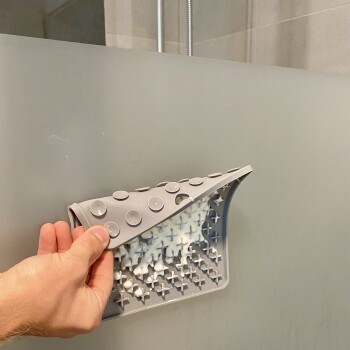 Eine Hand hält ein graues Silikon-Haustierpflegewerkzeug mit Saugnäpfen, das teilweise eine strukturierte Reinigungsoberfläche vor einem matten Hintergrund zeigt.