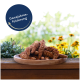Ein Holzbrett mit Vogelfutterstangen und einem blauen Etikett "Ganzjahresfütterung," umgeben von bunten Blumen und Grün.