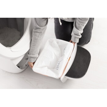 Weiße automatische Katzentoilette mit offener, herausziehbarer Schublade, ausgelegt mit Plastiktüte (gedruckter Text sichtbar) und schwarzer Riffelmatte.