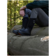 Eine Person, die auf einem Baumstamm im Wald sitzt, trägt schwarze Gummistiefel mit orangefarbenen Sohlen und graue Hosen, mit einem grünen Pullover.