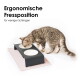Getigerte Katze frisst aus einem erhöhten schwarzen Doppelnapf mit weißen Schalen auf einer rosa Matte; sichtbarer Text: 'Ergonomische Fressposition'.