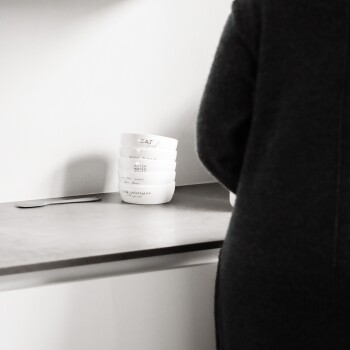 Eine Person in einem schwarzen Pullover steht in der Nähe einer Arbeitsplatte mit gestapelten weißen Futternäpfen, die mit "KATZE", "FUTTER BITTE" und "WASSER" beschriftet sind.