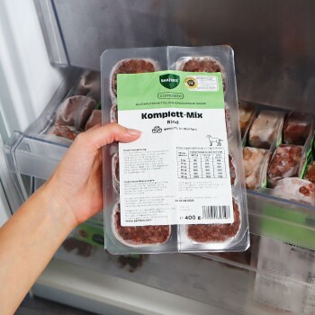 Hand hält verpackte Tiefkühlschale mit Hundefutter 'Komplett‑Mix Rind' (BARFER'S Komplett), 400 g, im Gefrierschrank.