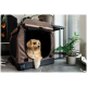 Ein kleiner Hund schaut aus einer stilvollen braunen Haustiertasche mit Netzpaneelen, die neben einer Topfpflanze in einem modernen Innenraum steht.