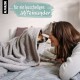 Ein Hund, eingewickelt in eine graue Decke, liegt auf einem Bett und schaut eine Person mit langen Haaren an. Textüberlagerung: '... für ein kuscheliges Miteinander.'
