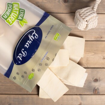 Eine Packung Lyra Nature Power Haustierleckerlis, mit rechteckigen Snacks, die auf einer Holzoberfläche verstreut sind, und einem Seilspielzeug im Hintergrund.