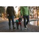 Eine Frau in einer grünen Jacke und ein Mann in einer grünen Steppjacke führen einen schwarzen Hund, der einen rot-grünen Mantel trägt, an einem Teich in einem Park.