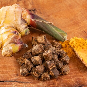 Eine Nahaufnahme von frischem Ingwerwurzel, Kurkumapulver und kleinen dunklen Tierfutterkibbles auf einer Holzoberfläche.
