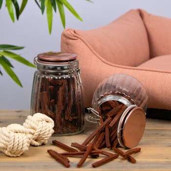 Zwei Glasbehälter mit Hundeleckerlis, einer umgekippt, der auf eine Holzoberfläche neben einem plüschigen Haustierbett und zwei Seilspielzeugen fällt.